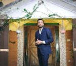 Man in a blue suit standing in front of a doorway, surrounded by greenery and hanging planters.