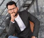 Confident man in a suit with eyeglasses and facial hair sitting indoors, exuding professionalism.